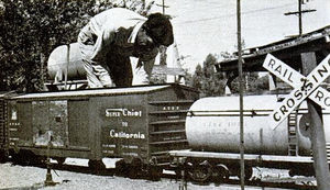 Mann gives appearance of a giant in Lilliputian land as he checks out freight car at railroad crossing.